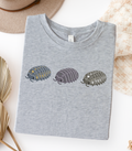 A folded light grey t-shirt with a graphic of three stylized isopods in blue and yellow, purple, and black and white.