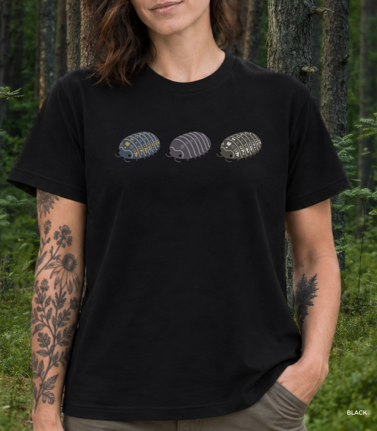 A women with floral tattoos standing in a forest wearing a black t-shirt with a graphic of three stylized isopods in blue and yellow, purple, and black and white.