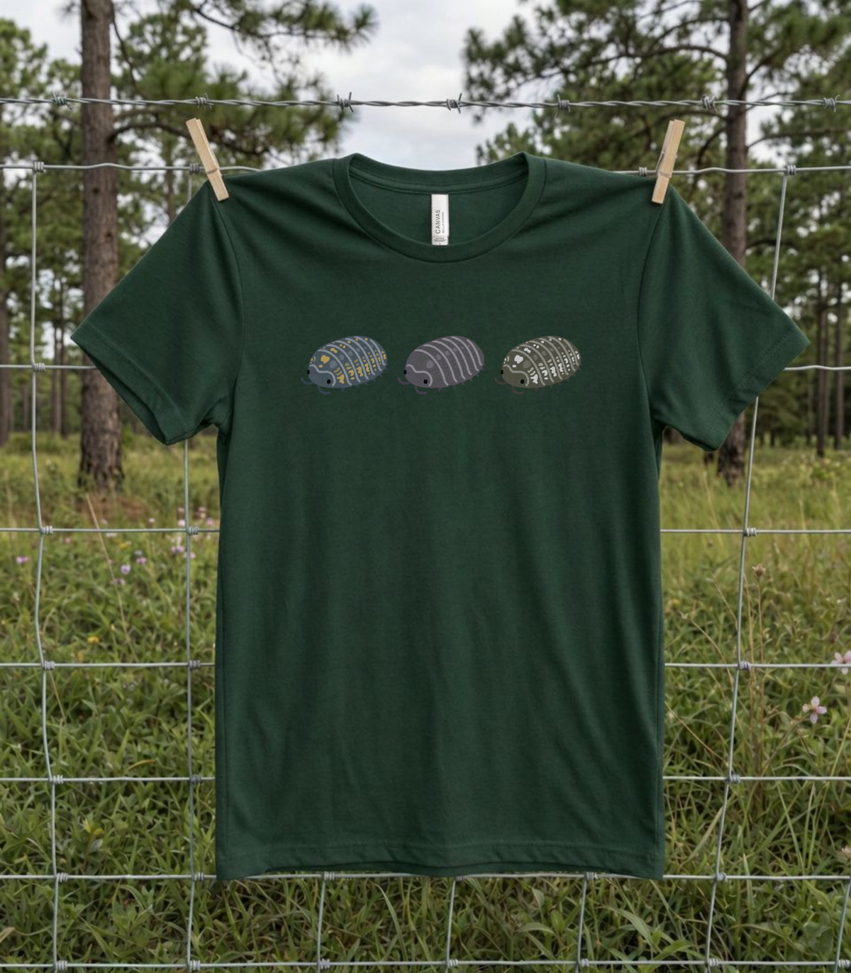 A forest green t-shirt hanging on a wire fence with a graphic of three stylized isopods in blue and yellow, purple, and black and white.