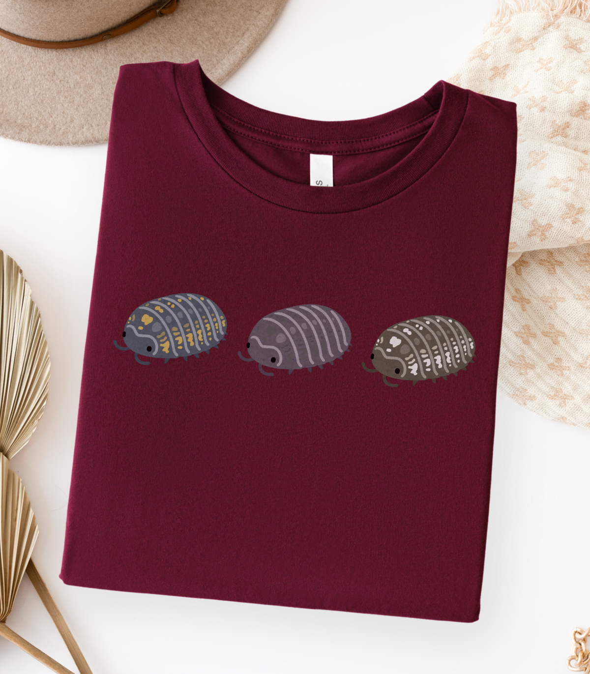 A folded maroon t-shirt with a graphic of three stylized isopods in blue and yellow, purple, and black and white.